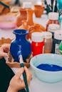 A person carefully paints a ceramic vase blue surrounded by vibrant paints and pottery.